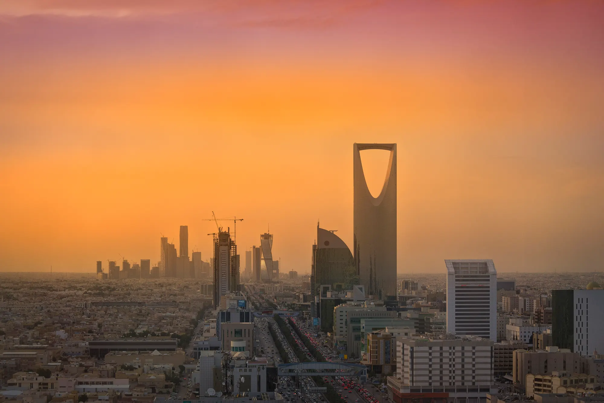 Riyadh at golden hour: the Kingdom Centre and cranes against an orange sky, the financial district beyond